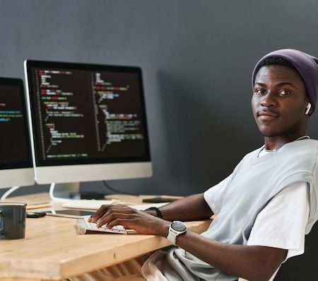 Young black man in casualwear keeping hands on computer keyboard while sitting in front of monitors _ Premium Photo